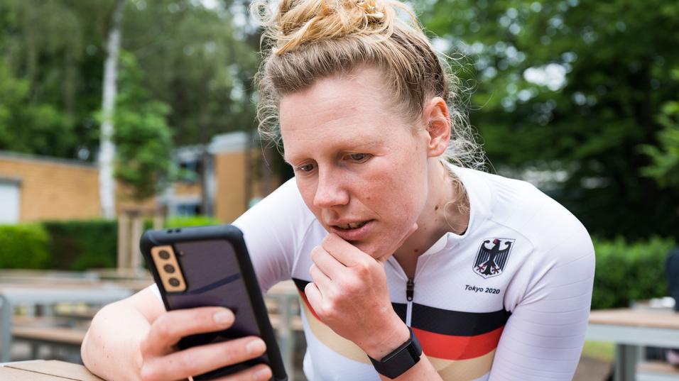 Athletin im weißen Trikot mit schwarzem und rotem Streifen schaut konzentriert auf ihr Smartphone in einer Außenanlage.